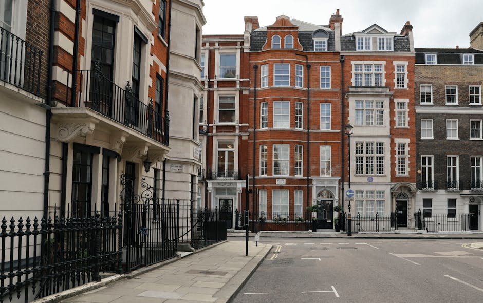 A street view in Belgravia showing a row of multi-storey residential buildings with traditional architecture, featuring brick facades, large sash windows, and ornate white decorative details around doors and balconies. In the foreground, a paved pavement with black wrought iron railings borders the buildings, and there is a black metal gate leading into the property. Parked along the street are several vehicles, including a white van positioned near one of the buildings, suggesting a home relocation or furniture transport activity. The scene is illuminated by daylight, with some equipment such as trolleys or moving blankets potentially present during a loading process conducted by Man and Van Belgravia for house removals or packing and moving services in the area.