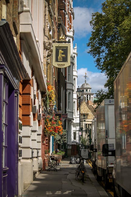 The image depicts a narrow street in Belgravia, lined with multi-storey Victorian and Georgian-style buildings featuring brick and stucco facades, decorative iron balconies, and flower boxes with vibrant plants. A white sign with gold lettering and an ornate emblem hangs from one building, indicating a pub or tavern. Parked on the street are several delivery trucks with reflective surfaces and black side mirrors, positioned close to the pavement. The foreground shows the side of a large white moving van, with an open side door for loading or unloading items, and adjacent to it, a few cardboard boxes, plastic-wrapped furniture, and packing materials are visible on the pavement. The scene suggests a home relocation or furniture transport process, with the loading area set up for a removal service by Man and Van Belgravia, amid the backdrop of the historic cityscape, including a domed church tower visible beyond the buildings, under a partly cloudy sky with dappled sunlight illuminating the scene.
