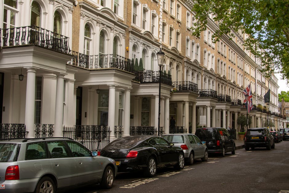 A street view in Belgravia showing a row of multi-storey residential buildings with traditional architecture, featuring brick facades, large sash windows, and ornate white decorative details around doors and balconies. In the foreground, a paved pavement with black wrought iron railings borders the buildings, and there is a black metal gate leading into the property. Parked along the street are several vehicles, including a white van positioned near one of the buildings, suggesting a home relocation or furniture transport activity. The scene is illuminated by daylight, with some equipment such as trolleys or moving blankets potentially present during a loading process conducted by Man and Van Belgravia for house removals or packing and moving services in the area.